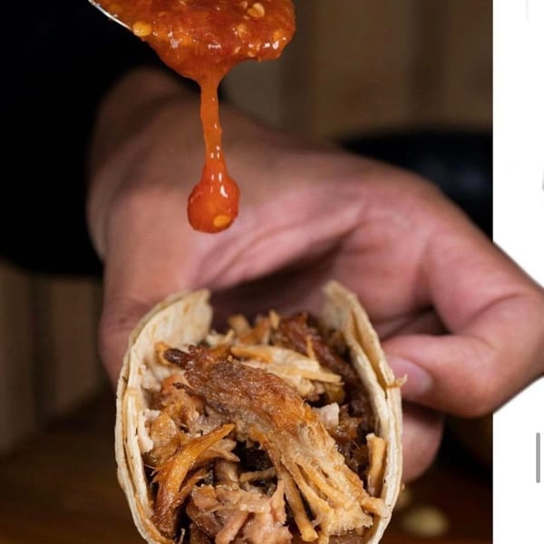 A hand holds a carnitas taco as red salsa drips from a spoon onto it.